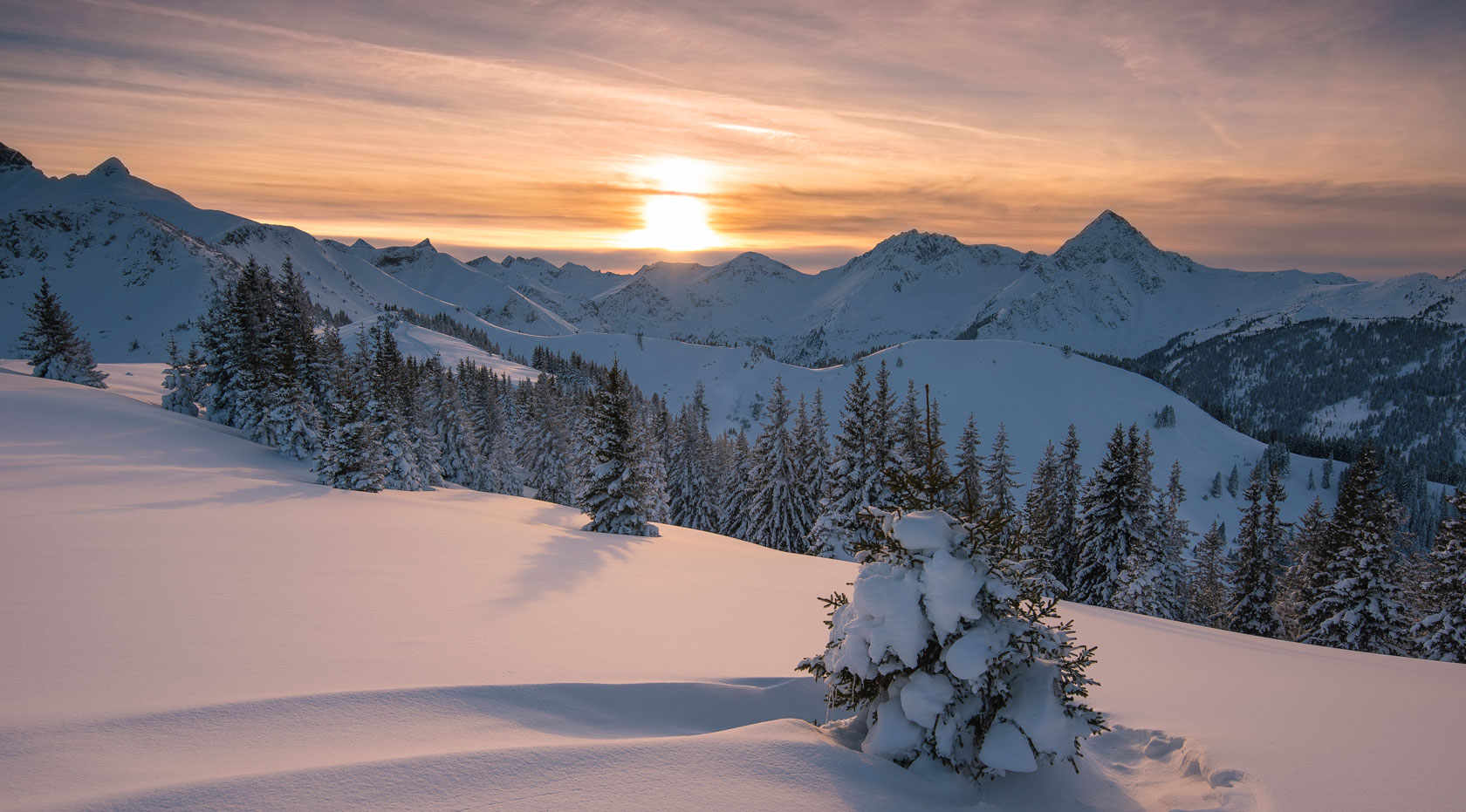 Verschneite Alpenlandschaft bei Sonnenuntergang mit tief verschneiten Tannen und Berghängen, warmes Licht im Winterpanorama.