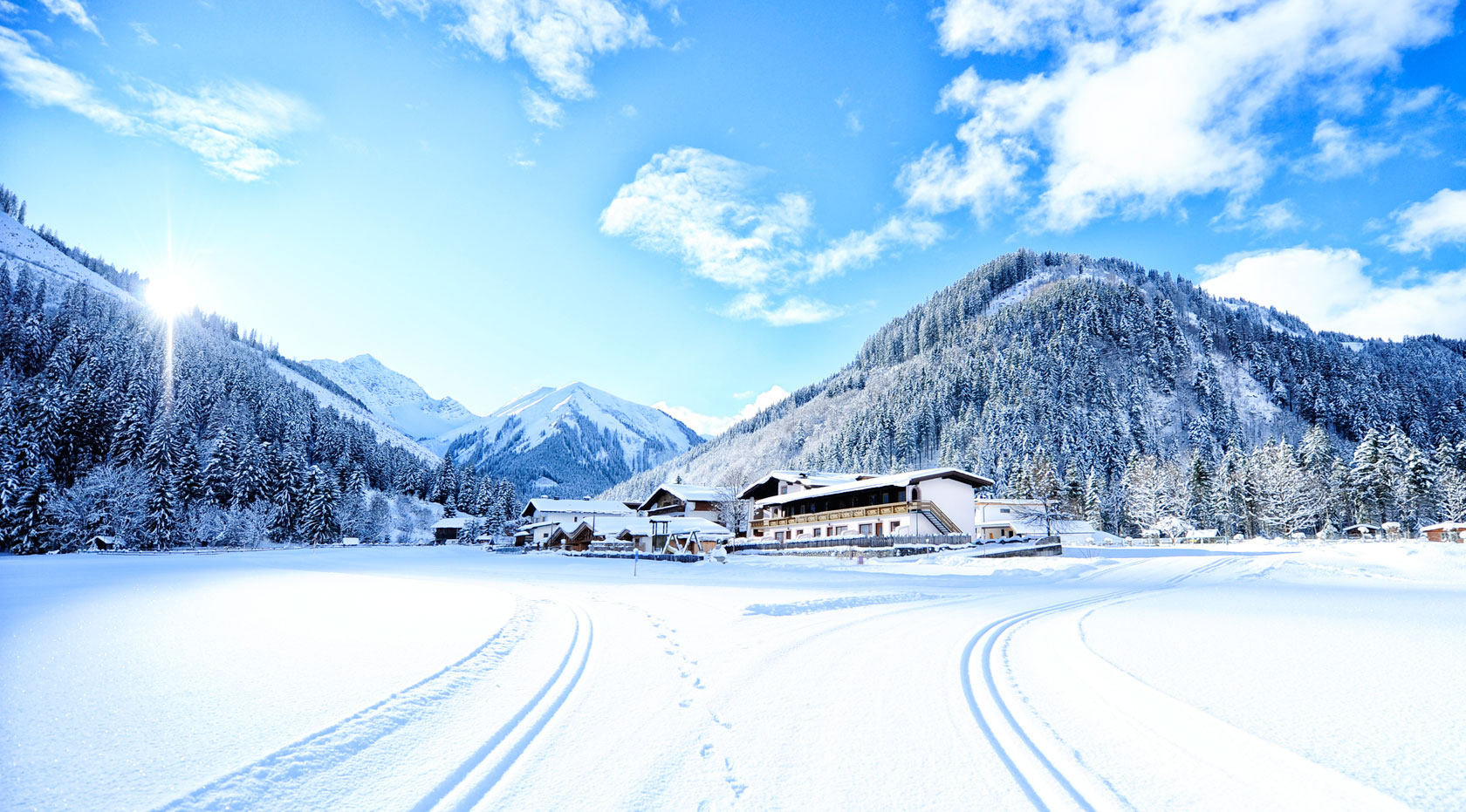 Verschneite Winterlandschaft mit Loipe, sonnigem Himmel, Chalets und bewaldeten Bergen in der Tiroler Alpenregion.