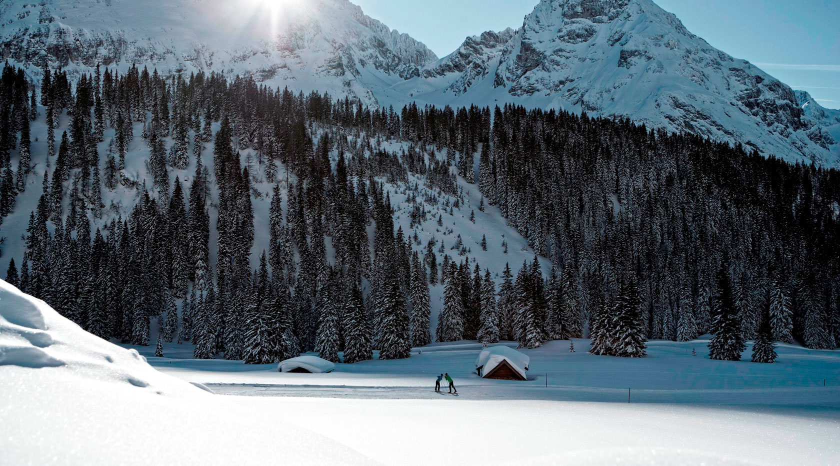 Zwei Langläufer in verschneiter Winterlandschaft mit tiefem Wald und imposanten Bergen im Hintergrund bei Sonnenschein.