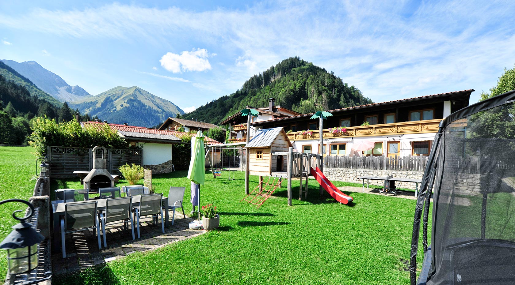 Der Spielplatz mit kleiner Holzhaus und Rutsche, sowie einem Trampolin und einem Tisch mit einigen Stühlen.