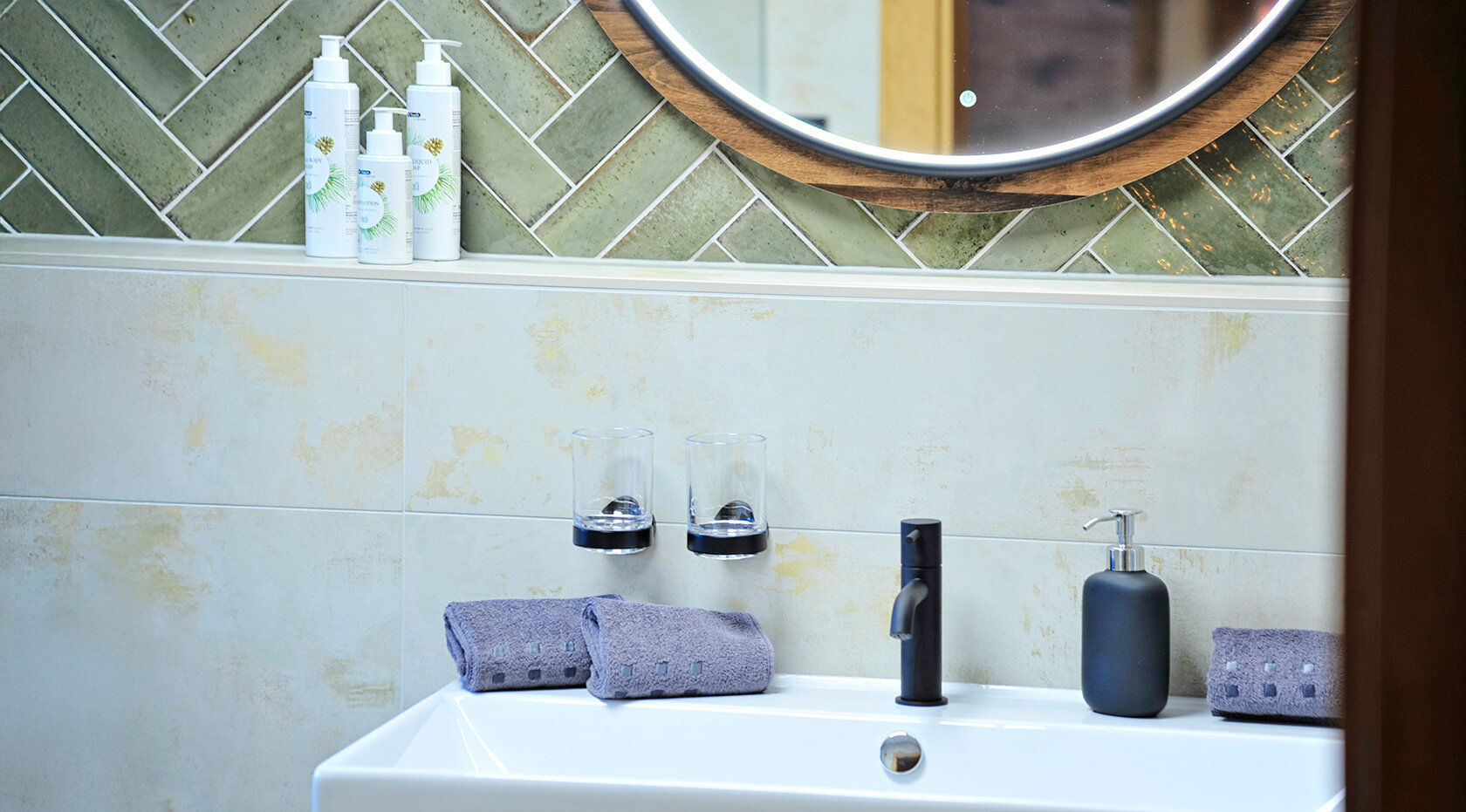 Modernes Badezimmer-Waschbecken mit zwei Armaturen, rundem Spiegel, Handtüchern und Seifenspendern vor heller Steinwand – Ferienwohnungen Wiesenruh.