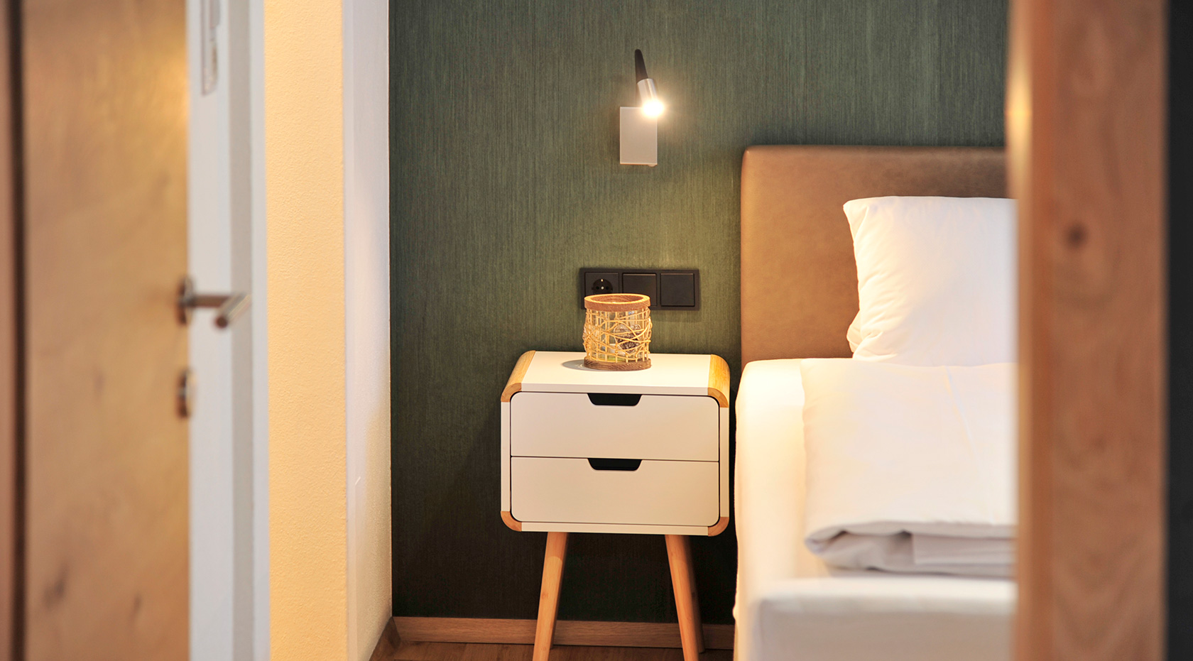 Schlafzimmer mit warmem Licht: Blick durch Holztür auf Bett, Nachttisch mit Lampe und Deko vor dunkler Akzentwand – Ferienwohnungen Wiesenruh.