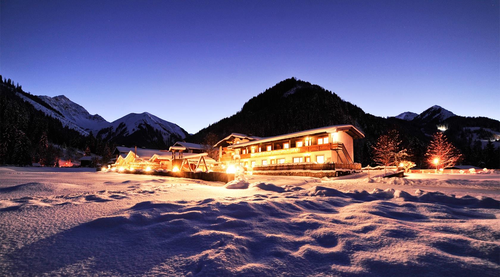 Verschneite Chalets in den Alpen bei Dämmerung, warm erleuchtet vor winterlicher Bergkulisse.